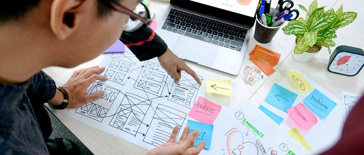 A lot of UX design documents on a table plus a laptop, 4 hands from 3 different people and part of a face looking at the documents