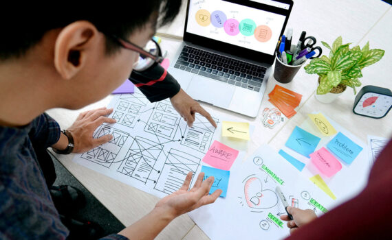 A lot of UX design documents on a table plus a laptop, 4 hands from 3 different people and part of a face looking at the documents