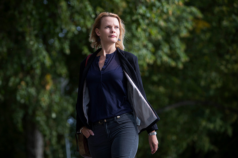 A blond woman walking towards the camera wearing a black jacket and dark blouse, with green trees in the background